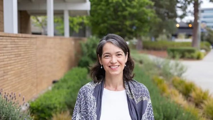 Rebecca Delafield stands outdoors on a campus walkway, smiling toward the camera, with landscaping, buildings and a path receding behind her.