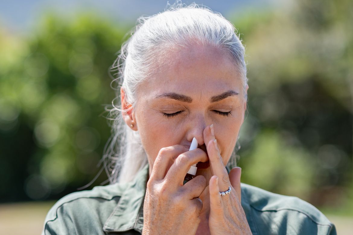Woman with grey hair closes left nostril with hand and uses white nasal spray bottle in right nostril