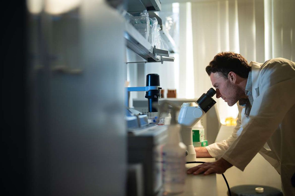 David Nathanson in a white lab coat looks through a microscope at a laboratory bench stocked with scientific equipment and supplies.