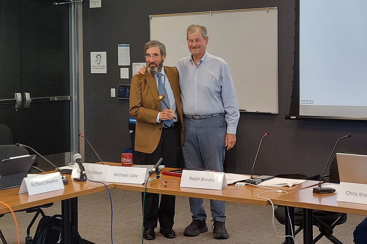 Richard Shemin, left, and Ralph Brindis pose together behind a conference table; Shemin holds a small gavel while standing in front of nameplates.