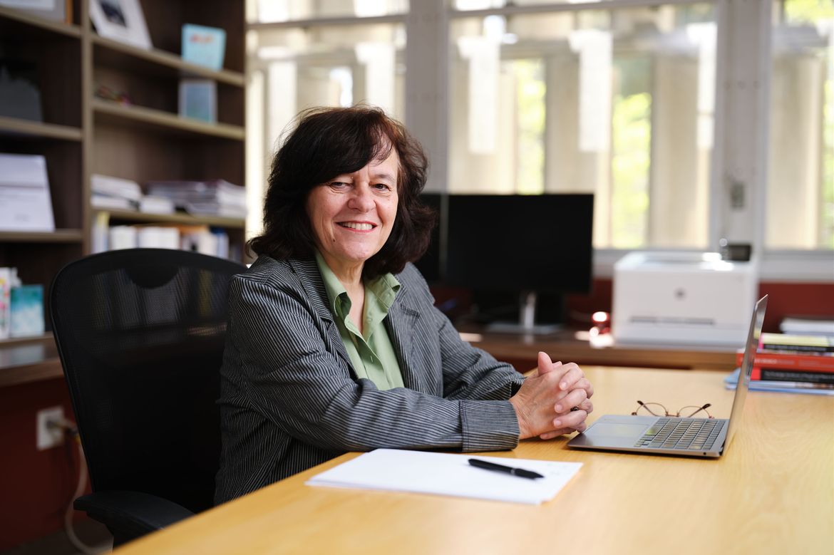 Anastasia Loukaitou-Sideris, in green top and striped jacket, sitting at her desk with hands clasped in front