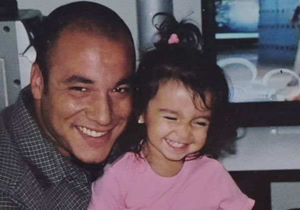 A smiling man poses beside a young girl in a pink shirt as they sit together indoors.