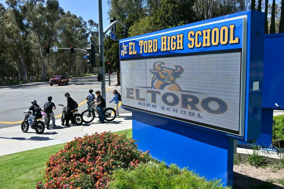 A sign for El Toro High School stands near a street corner as several students with e-bicycles gather at the intersection nearby.