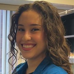 Clarissa Carillo, smiling warmly, with curly, shoulder-length hair, wearing a teal top, photographed indoors.