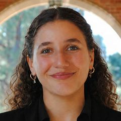 Gianna Karkafi, smiling, with curly, shoulder-length dark hair, wearing small gold hoop earrings and a dark top, photographed near a stone archway.