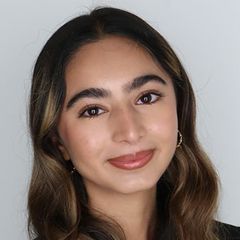 Sabah Alidina, smiling softly, with wavy, highlighted hair, wearing small hoop earrings and a dark top, against a light gray background.