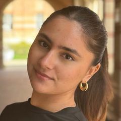 Tamanna Tikoo, with dark hair pulled back in a ponytail, wearing small gold hoop earrings and a dark top, photographed outdoors near a brick archway.