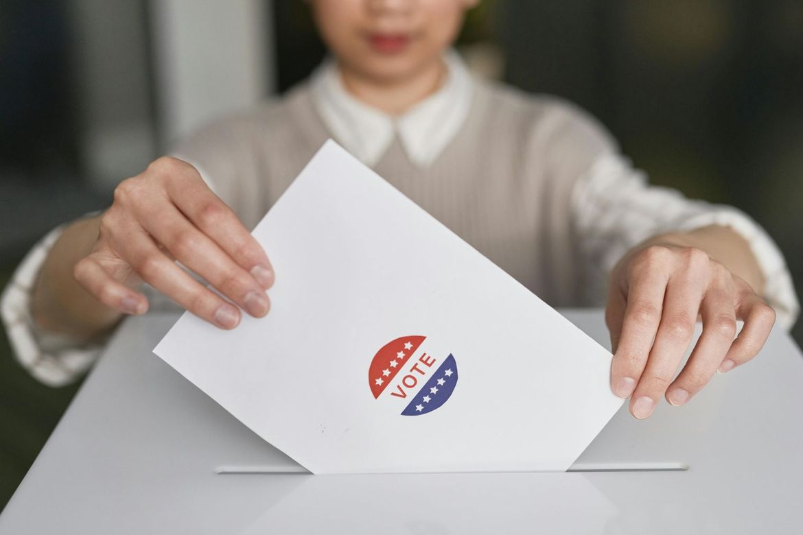 Woman drops a ballot envelope marked with a red white and blue Vote sticker into a cardboard ballot box