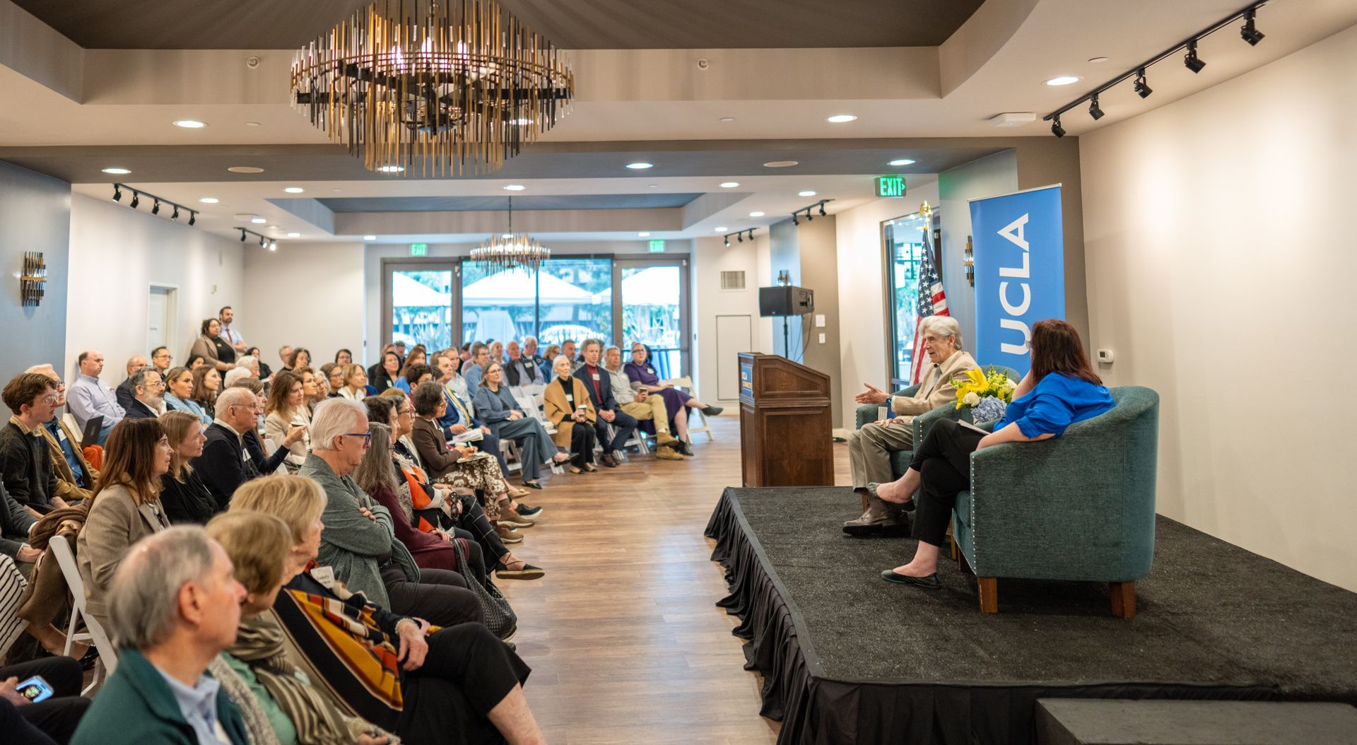 UCLA Chancellor Julio Frenk speaks during a community discussion at Belmont Village as audience members listen from rows of chairs facing a small stage with UCLA signage.