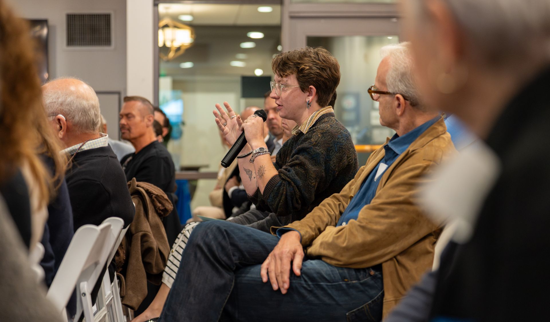 Everest Harvey speaks into a handheld microphone during a community discussion as attendees seated nearby listen inside a meeting room at Belmont Village.