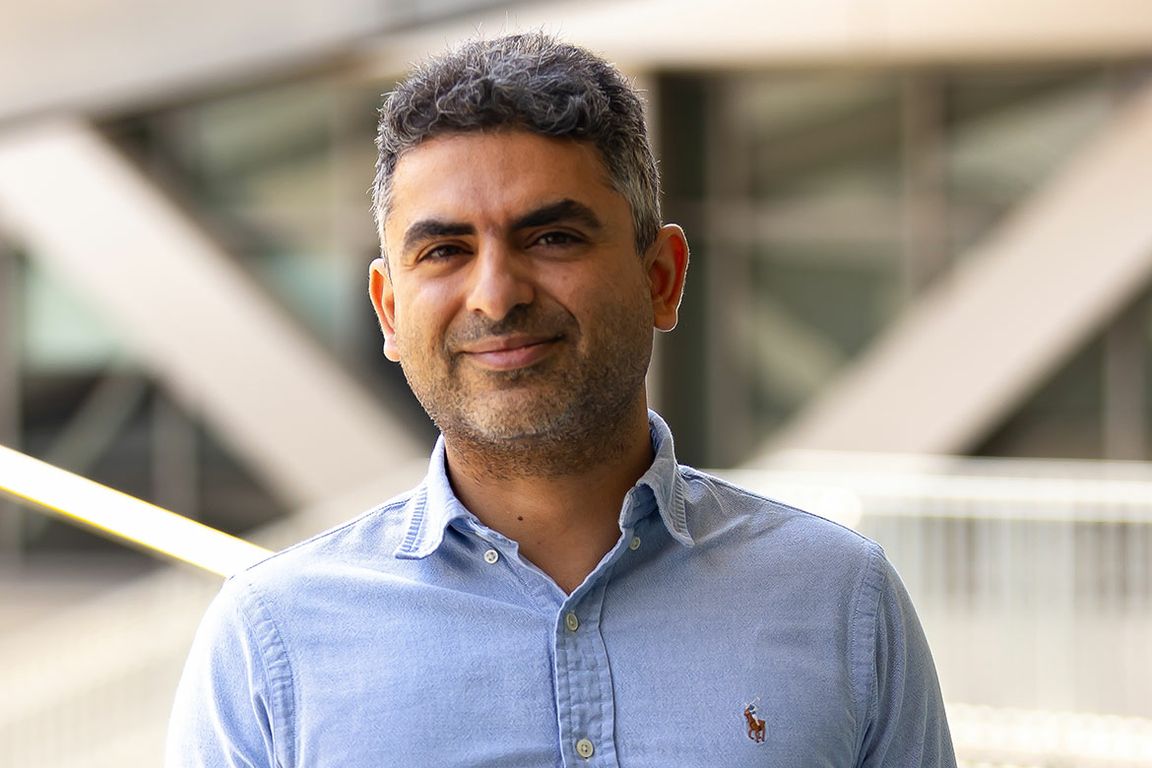 Headshot of Hosein Mohimani wearing a light blue button-down shirt, smiling in front of a modern building exterior.