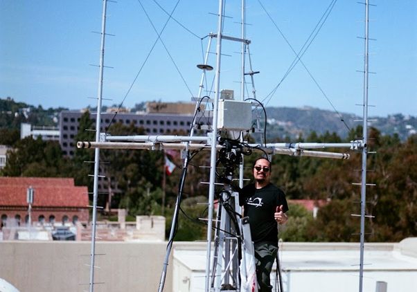 Emmanuel Masongsong in a black t-shirt stands on a rooftop surrounded by a large UHF antenna structure made of metal poles and cables, with the UCLA campus buildings and tree-covered hills visible in the background under a clear blue sky.