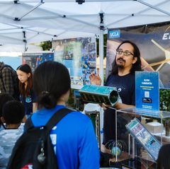 Emmanuel Masongsong, with long dark hair and glasses, holds a small rectangular satellite model at a UCLA booth under a white tent during the Exploring Your Universe 2023 event.