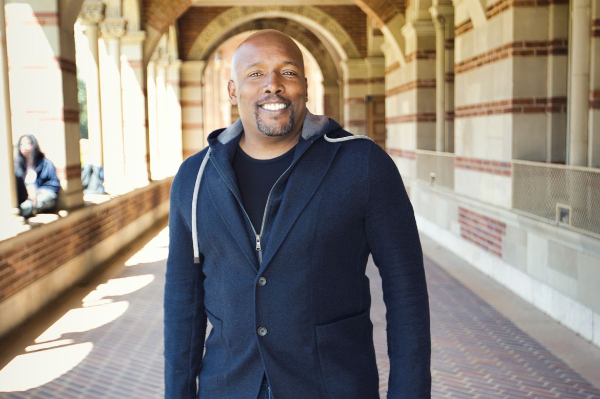 Kwame J. Granderson in a blue sweater, standing in the portico of Royce Hall on a sunny day.
