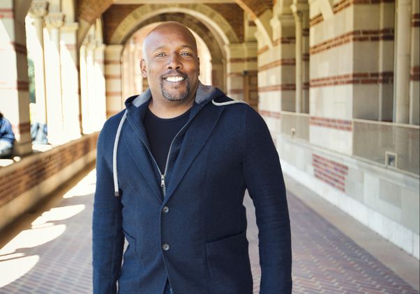 Kwame J. Granderson in a blue sweater, standing in the portico of Royce Hall on a sunny day.