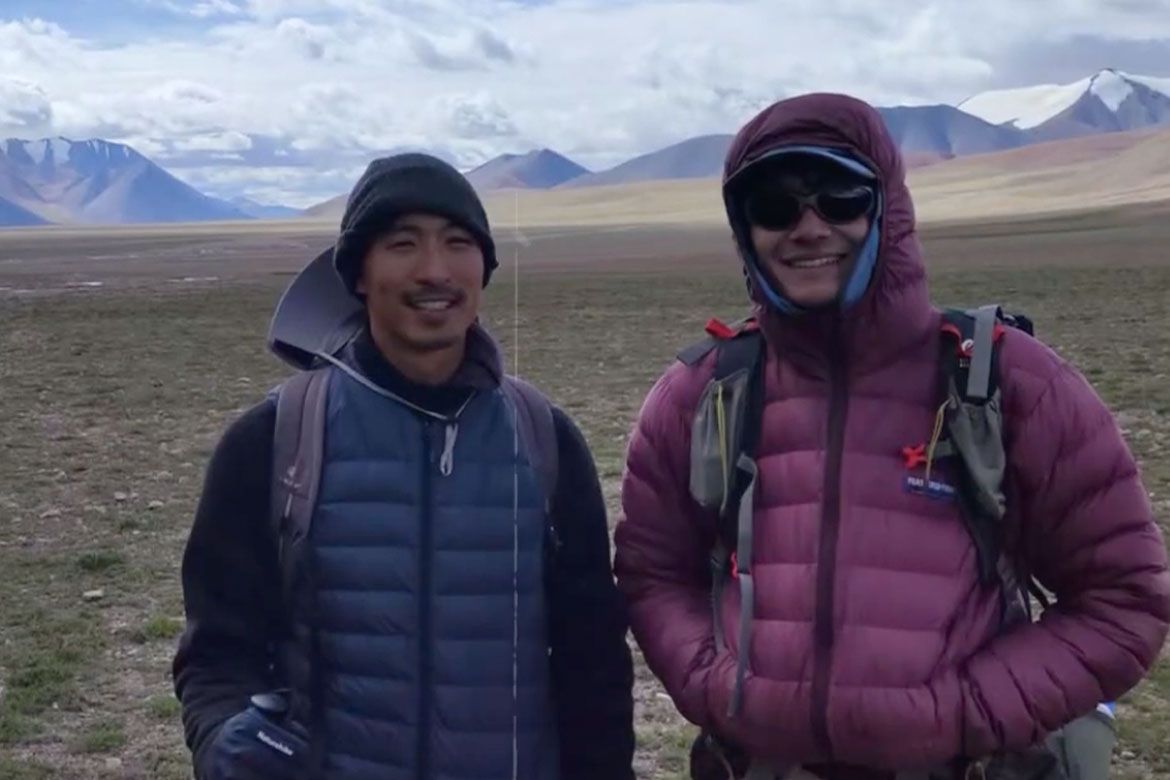 John He and Jordan Wang smile for a photo while conducting fieldwork in Tibet's Qiangtang Wilderness Preserve, with a vast high-altitude plateau and snow-capped mountains behind them.
