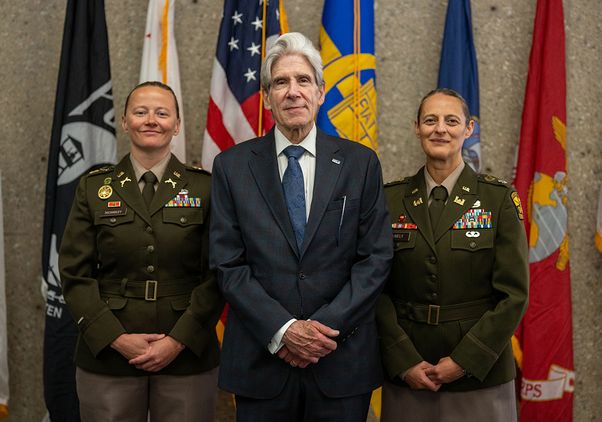 Chancellor Julio Frenk, wearing a dark navy suit and a blue-patterned tie, stands in the center between two U.S. Army officers in olive-green dress uniforms adorned with medals and ribbons. 