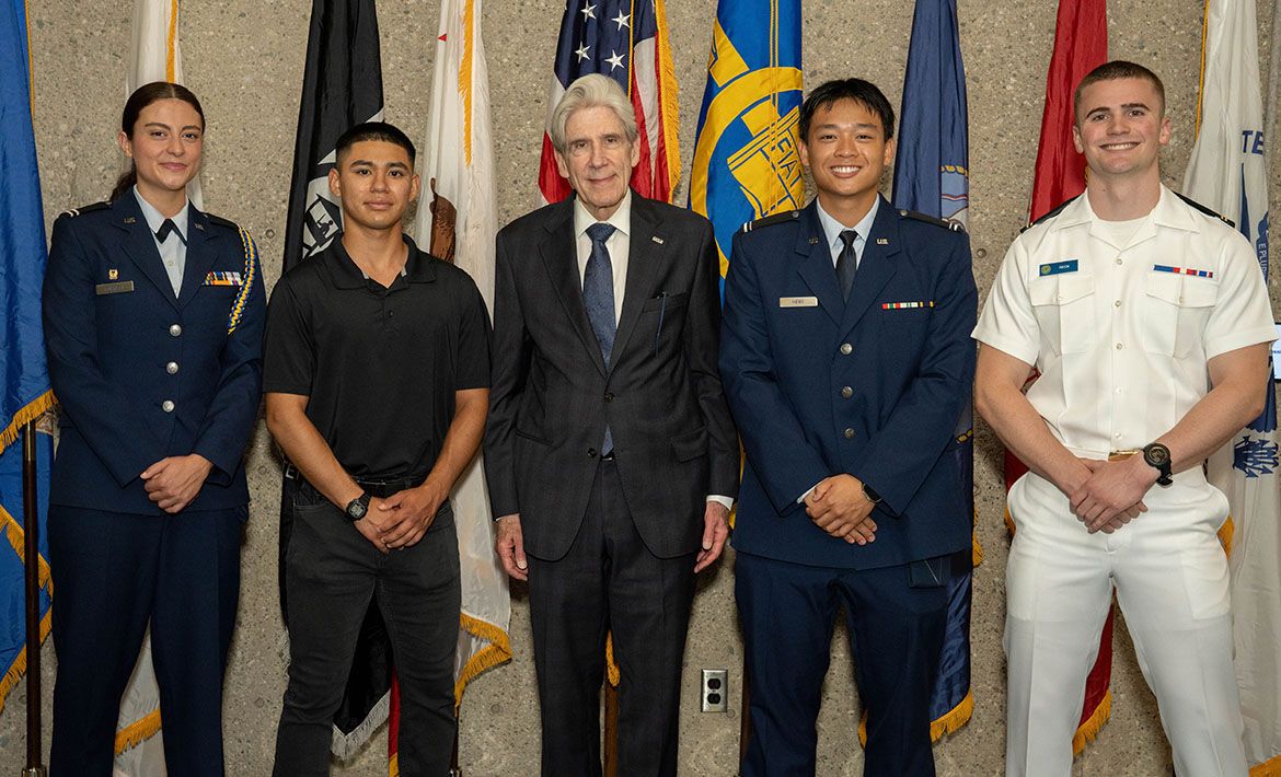 Chancellor Julio Frenk poses with four students and service members in a mix of Air Force dress uniforms and Navy whites, standing before a display of military and institutional flags. L-R: Cadet Ashley Forrest (Air Force ROTC), Cadet Albert Picon (Army ROTC), Chancellor Julio Frenk, Cadet Eric Heng (Air Force ROTC), and Midshipman Nicolas Reck (Naval ROTC).