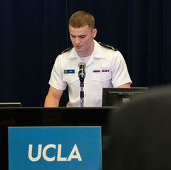 A military cadet in white uniform speaks at a UCLA-branded podium during a 