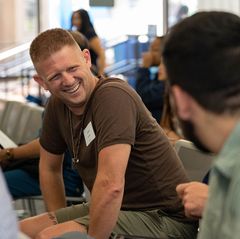 A man in a brown t-shirt laughs while seated among other attendees at a UCLA Memorial Day ceremony.