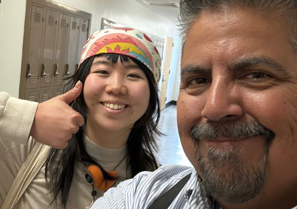 UCLA student Cylin Wang smiles and gives a thumbs-up while posing for a selfie with UCLA senior custodian Daniel Estrada in a campus hallway.