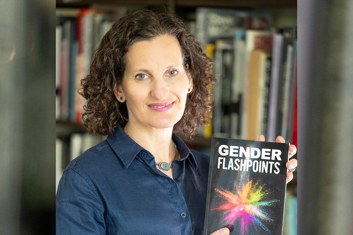 Abigail Saguy holds up a book titled "Gender Flashpoints," with a colorful powder burst on the cover. She stands in front of a bookshelf and is wearing a blue, button-down shirt.