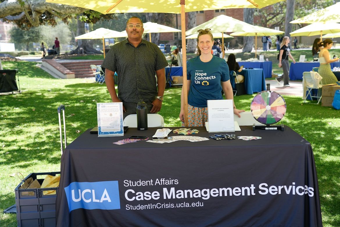 Two UCLA staff members stand behind a Student Affairs Case Management Services table during the Day of Hope mental health event, sharing wellness resources and student support information beneath umbrellas on the UCLA campus lawn.