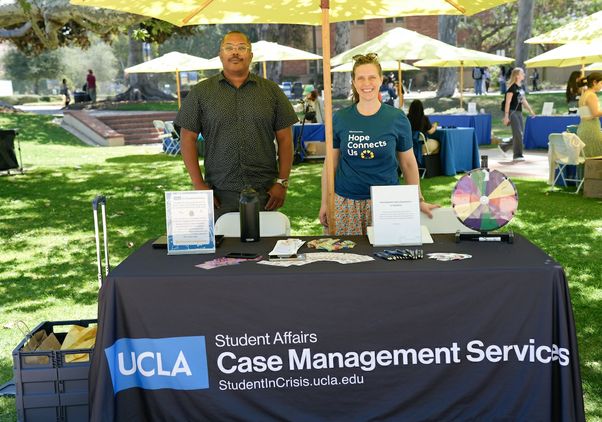 Two UCLA staff members stand behind a Student Affairs Case Management Services table during the Day of Hope mental health event, sharing wellness resources and student support information beneath umbrellas on the UCLA campus lawn.