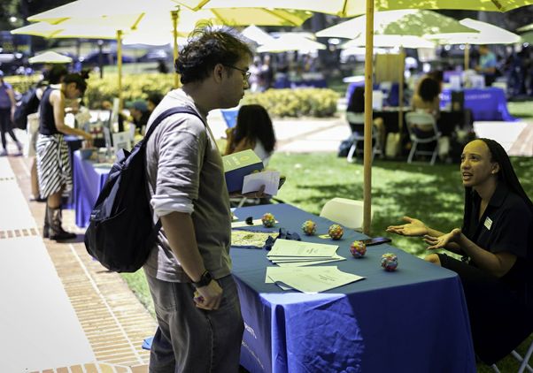 A UCLA student speaks with an event volunteer at an information table during the Day of Hope mental health event, exploring campus wellness resources and student support services beneath shaded umbrellas on campus.