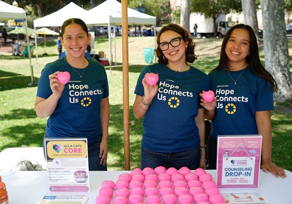 Three UCLA student volunteers wearing “Hope Connects Us” shirts smile behind a UCLA CAPS CORE table during the Day of Hope mental health event, sharing counseling and wellness resources with students on campus.