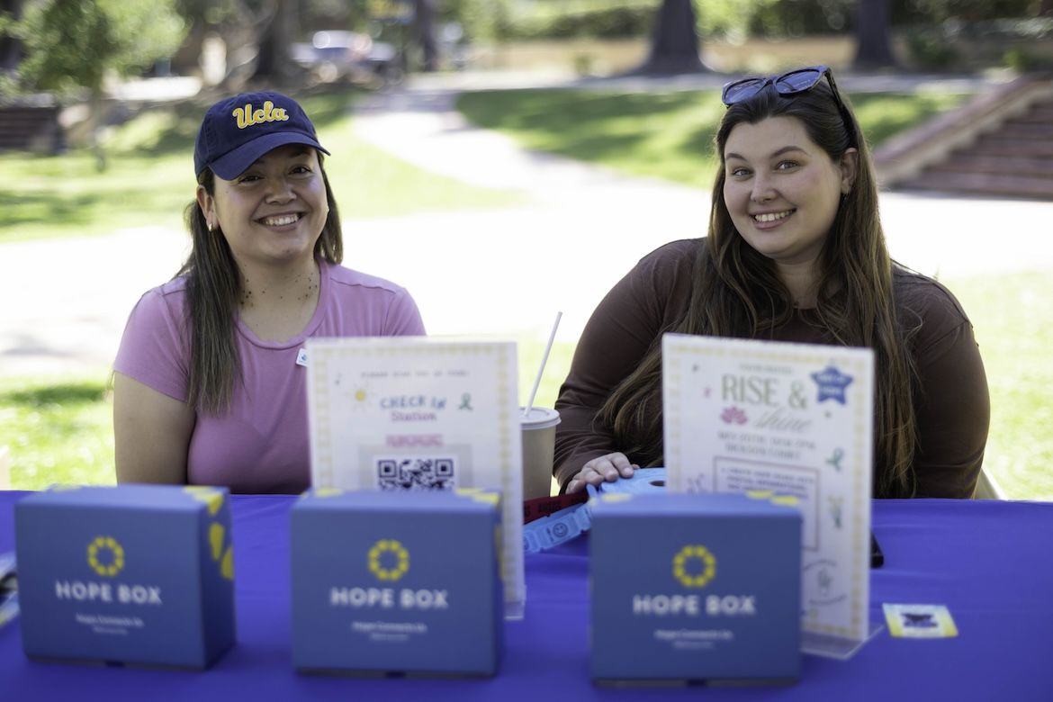 Two UCLA students smile behind a wellness resource table featuring Hope Boxes during the Day of Hope mental health event, helping promote emotional support, suicide prevention awareness and student connection on the UCLA campus.