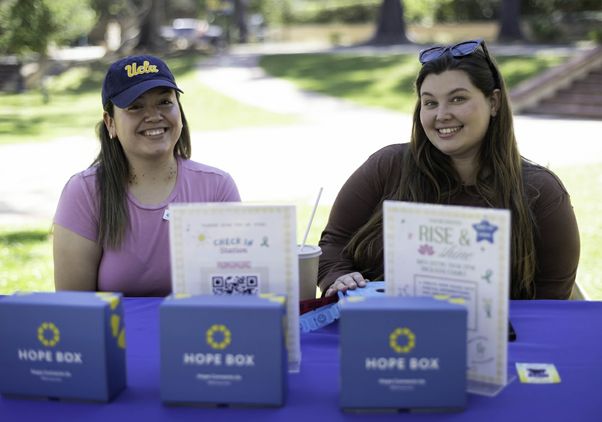 Two UCLA students smile behind a wellness resource table featuring Hope Boxes during the Day of Hope mental health event, helping promote emotional support, suicide prevention awareness and student connection on the UCLA campus.