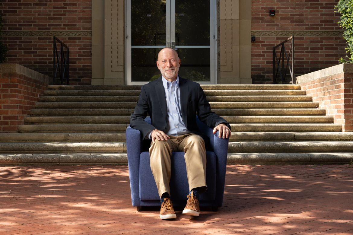 Shalom Staub in light-blue button-down, dark jacket and tan pants, seated in a blue armchair in front of a brick building