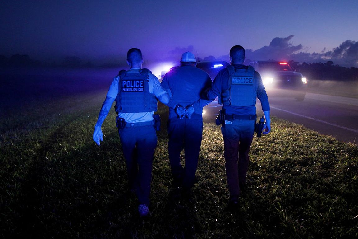 Two federal police officers in tactical vests escort a handcuffed individual along a roadside at night, illuminated by blue and red emergency lights from nearby vehicles.