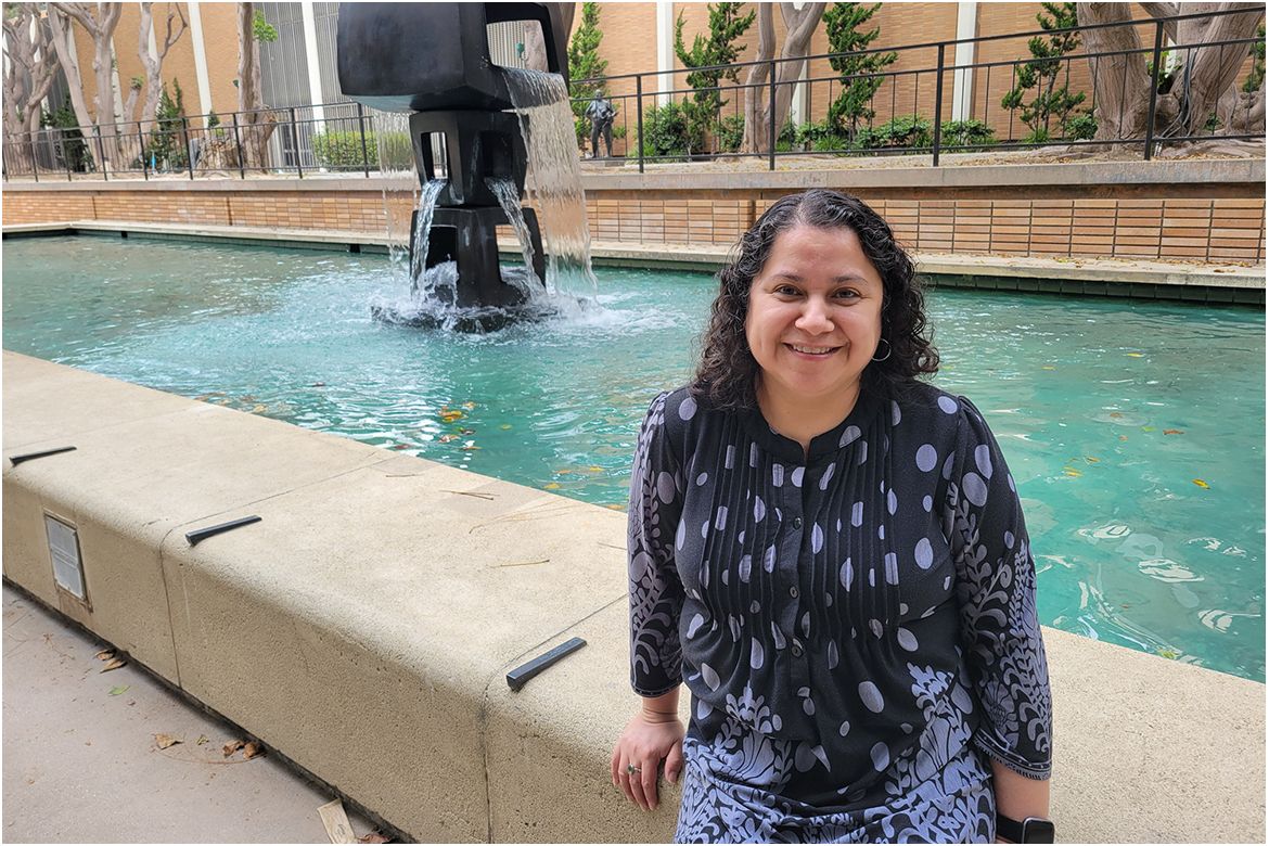 Leisy Abrego, in black and purple dress, sits on the edge of a rectangular outdoors water fountain.