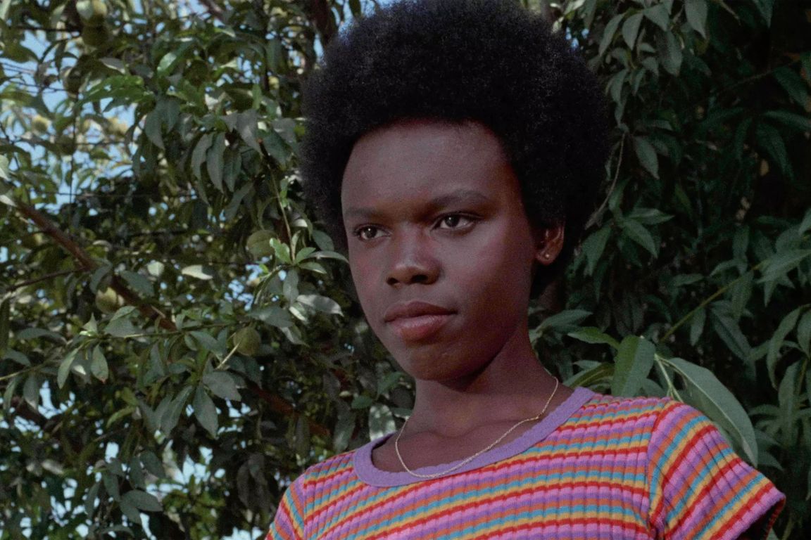 Peggy Pettitt, wearing a colorful striped shirt and a thin necklace, stands outdoors in front of leafy trees in a scene from the 1972 film “Black Girl.” 