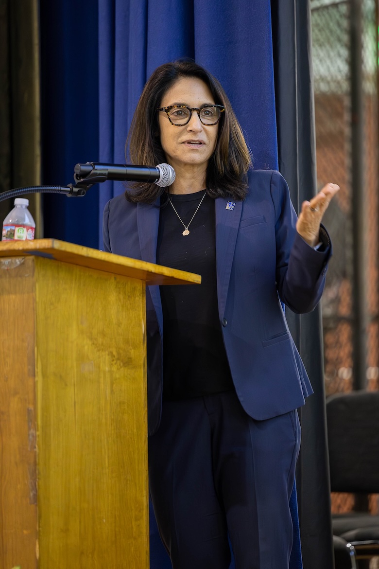 Christina Christie stands at a podium, speaking into a microphone and gesturing with one hand. She wears glasses and a navy suit, with a blue curtain behind her