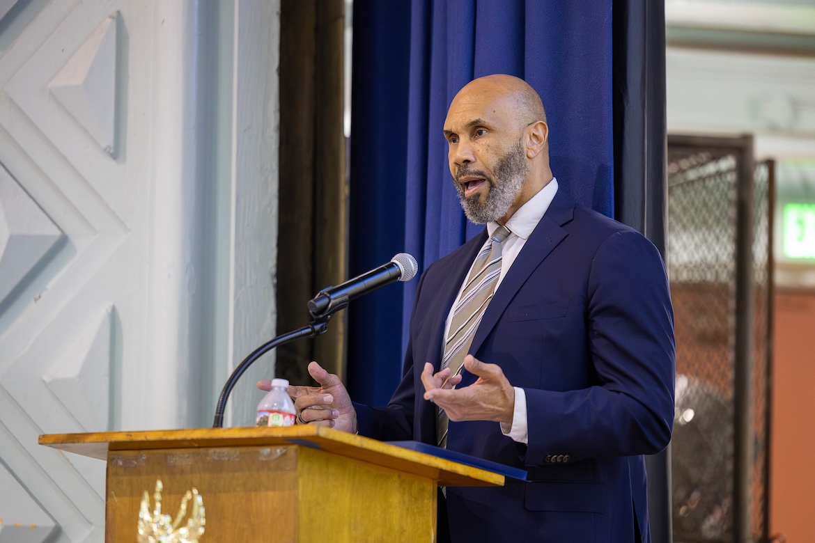 Darnell Hunt stands at a podium, speaking into a microphone while gesturing with his hands. He wears a dark suit and tie, with a blue curtain behind him