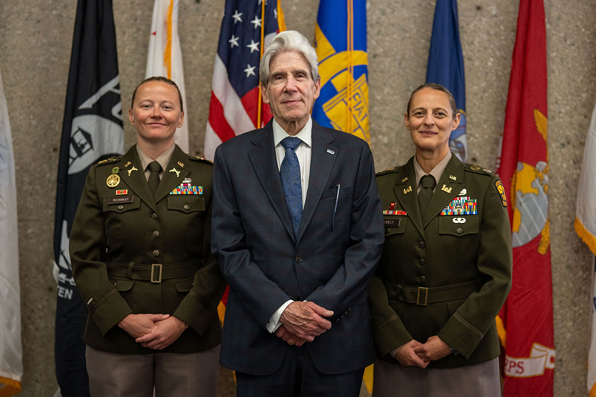 Chancellor Julio Frenk, wearing a dark navy suit and a blue-patterned tie, stands in the center between two U.S. Army officers in olive-green dress uniforms adorned with medals and ribbons. 