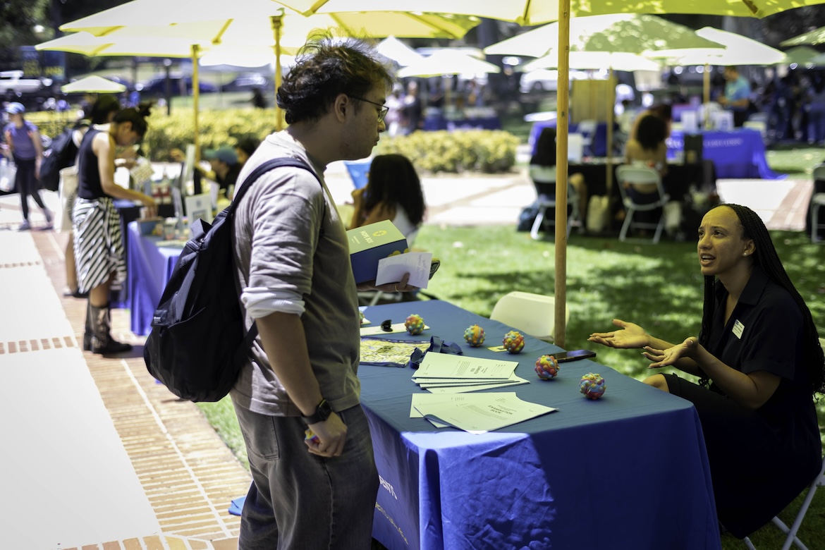  A UCLA student speaks with an event volunteer at an information table during the Day of Hope mental health event, exploring campus wellness resources and student support services beneath shaded umbrellas on campus.