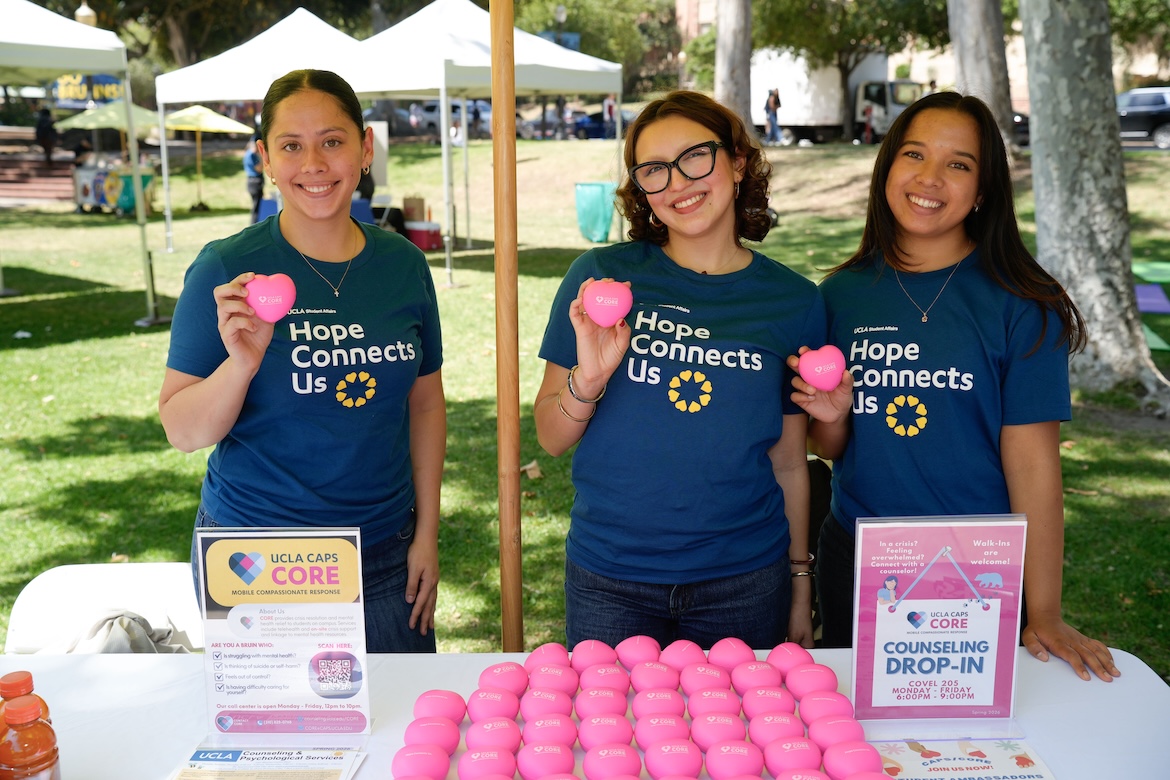 Three UCLA student volunteers wearing “Hope Connects Us” shirts smile behind a UCLA CAPS CORE table during the Day of Hope mental health event, sharing counseling and wellness resources with students on campus.