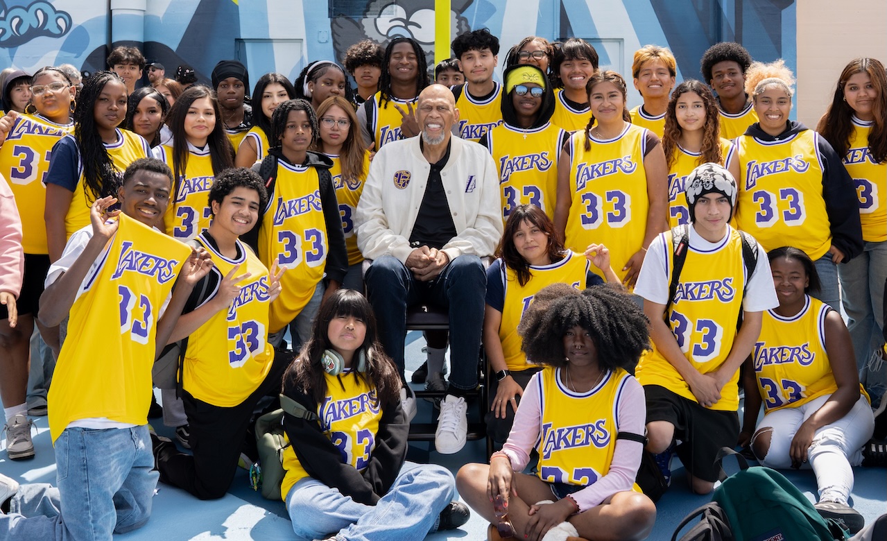 A group of Mann School students pose outdoors around an Kareem Abdul-Jabbar, who is seated at center — all students are wearing yellow Los Angeles Lakers jerseys with No. 33