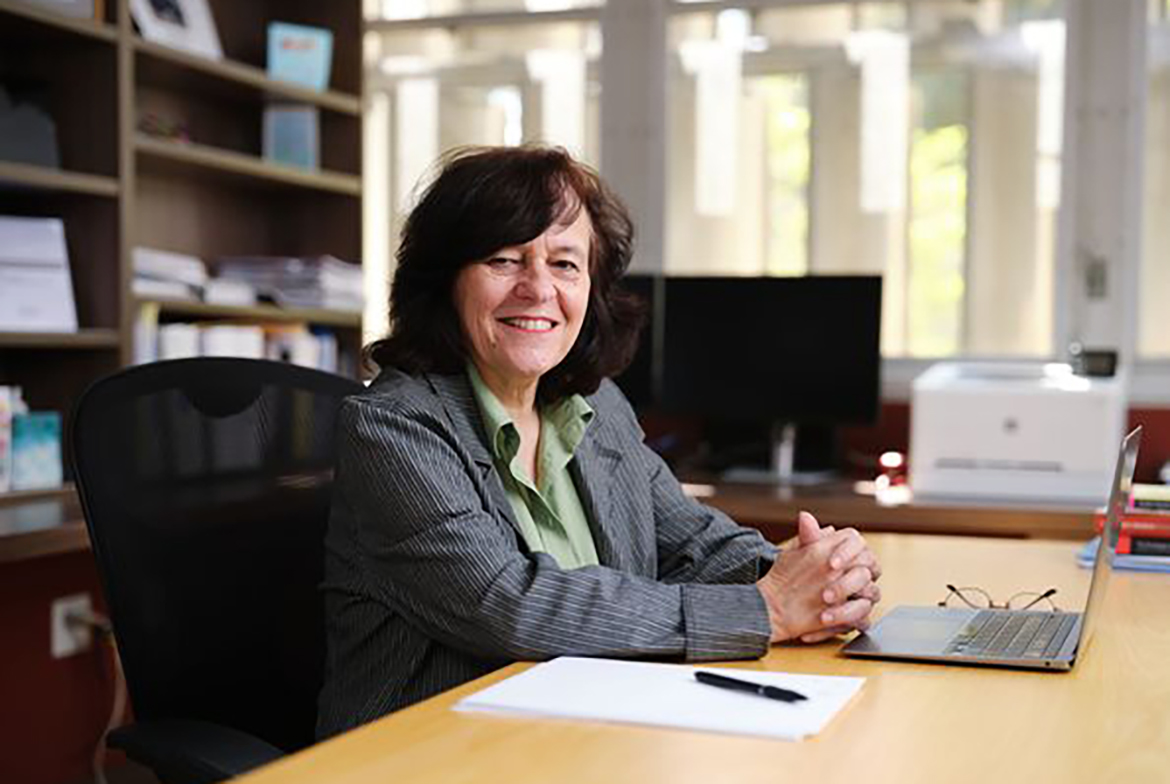 Anastasia Loukaitou-Sideris, in green top and striped jacket, sitting at her desk with hands clasped in front
