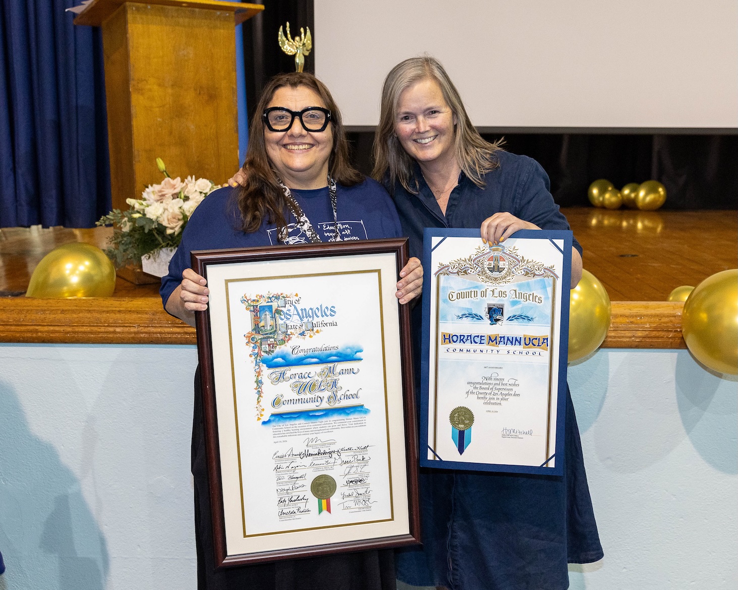 Mann UCLA principal Maria Cristina Phillips and UCLA’s Karen Hunter Quartz stand indoors, smiling and holding framed certificates from the city and county of Los Angeles recognizing Horace Mann UCLA Community School. 