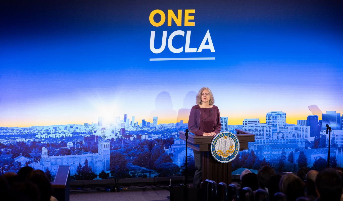 Megan McEvoy in plum top stands behind podium with UCLA seal and blue, white and yellow UCLA campus backdrop during 2026 State of the Campus address