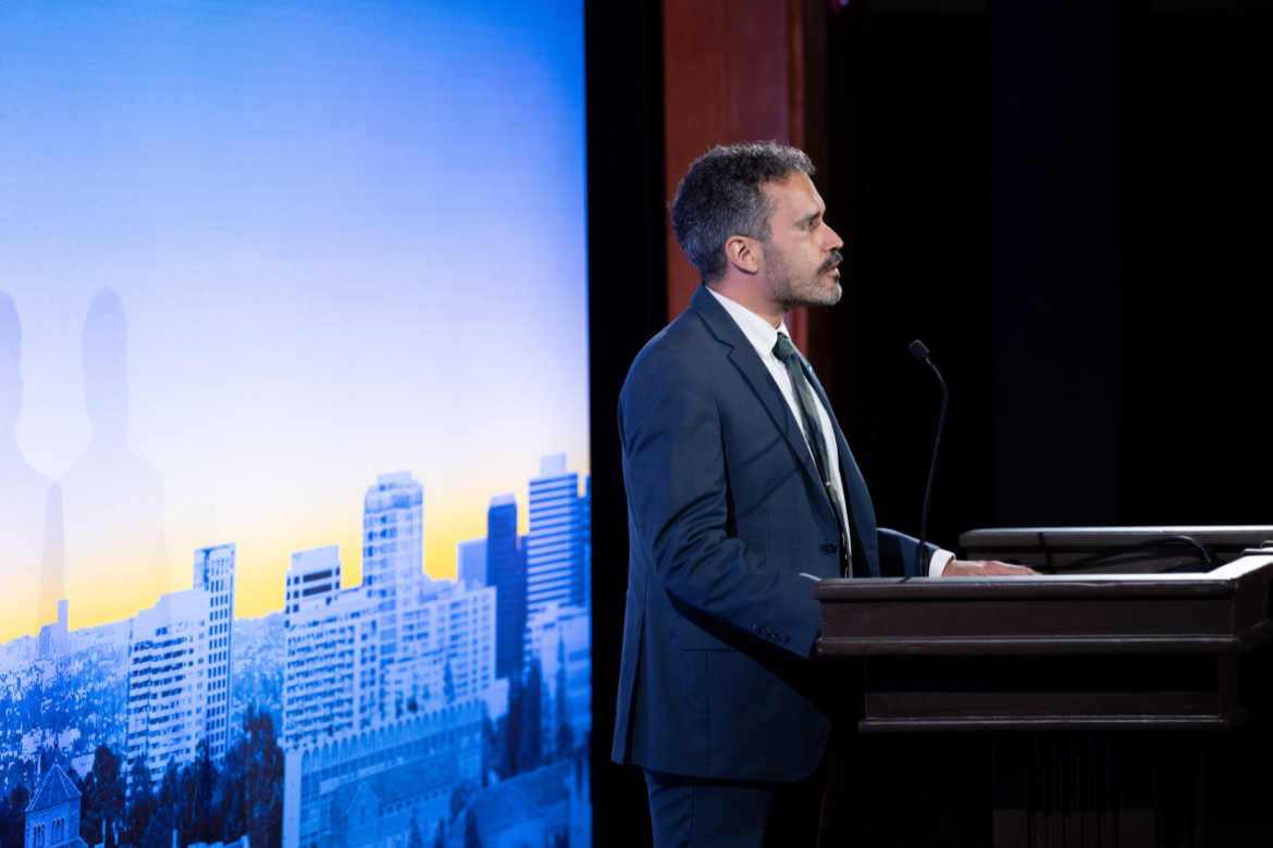 Michael McCormack-Colón in dark blue suit stands with hands on podium in front of blue, white and yellow UCLA campus backdrop during 2026 State of the Campus address
