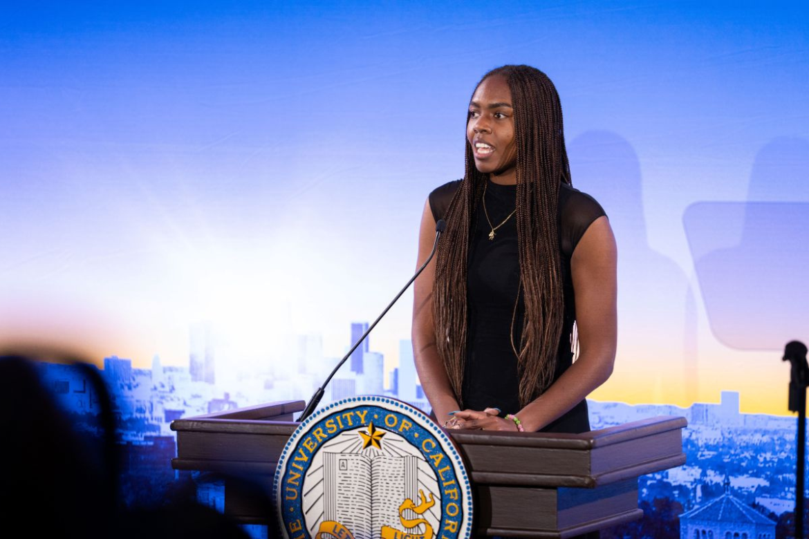Naomi Hammonds in black dress stands behind podium with UCLA seal and blue UCLA campus backdrop during 2026 State of the Campus address