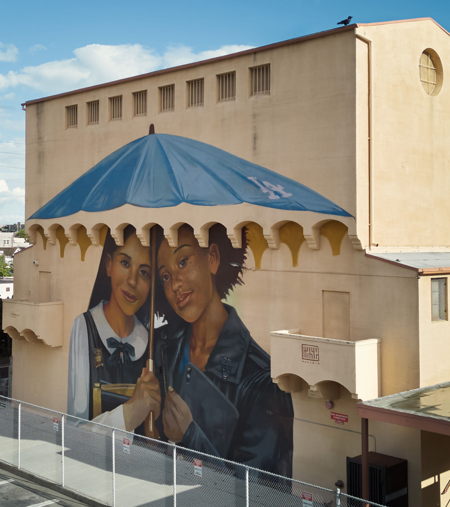 A large mural on a beige building shows two girls standing close under a blue umbrella marked with an “LA” logo. A chain-link fence runs in front.