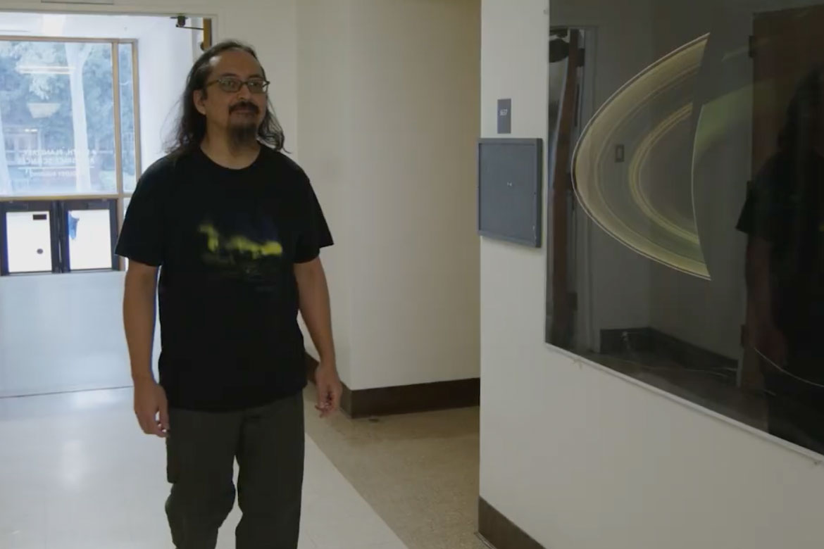 Emmanuel Masongsong, with long dark hair, glasses, and a black graphic t-shirt, walks through a university hallway. A large framed image of Saturn's rings is displayed on the wall to his right.