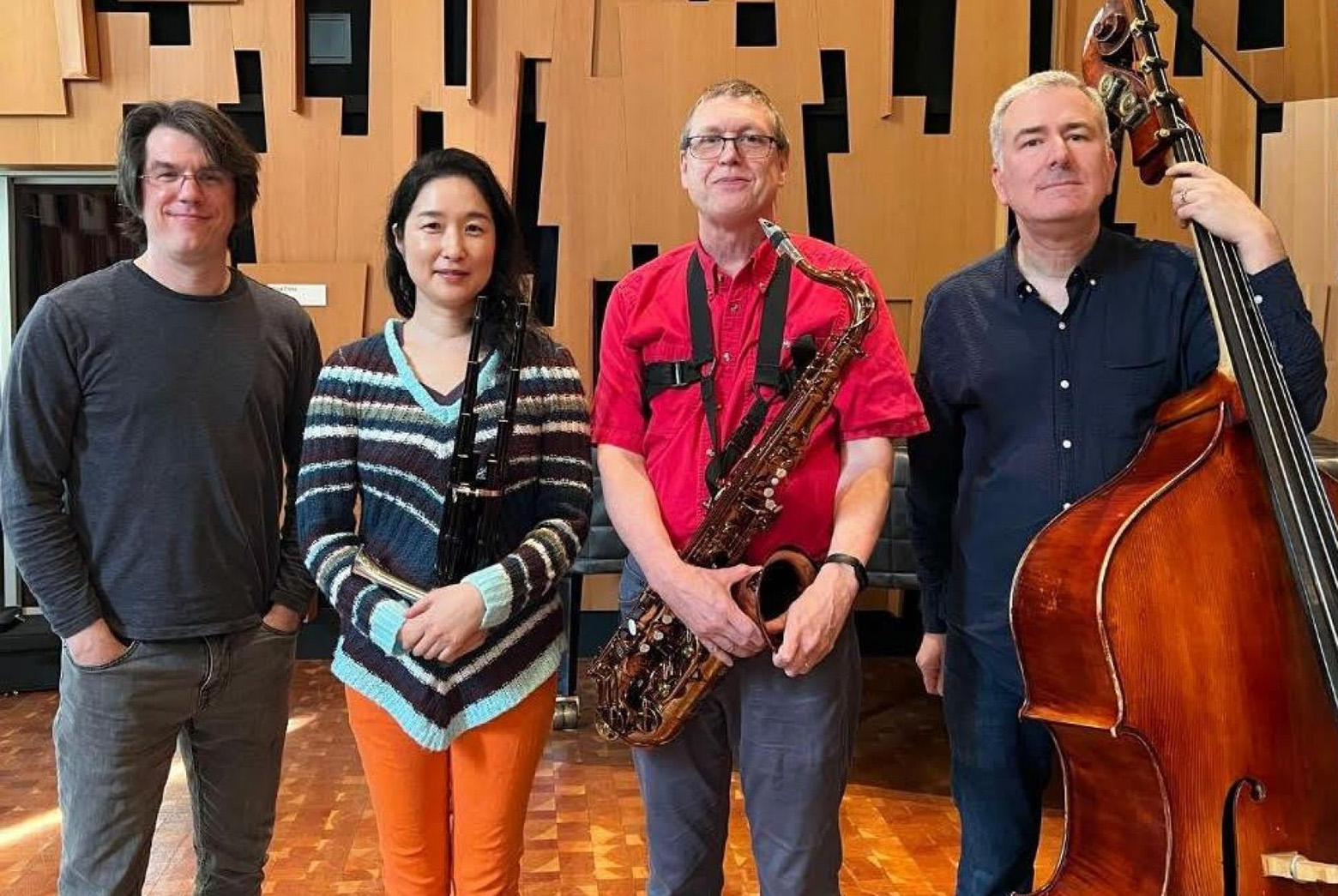 Four people, three holding instruments, pose for a photo; Robert Reigle, wearing a red shirt and holding a saxophone, is third from the left.